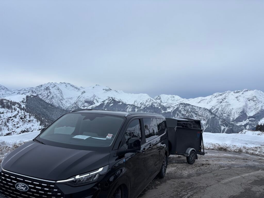 Van VTC noir avec remorque, garé en montagne face aux sommets enneigés des Alpes